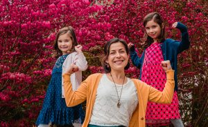 a woman in orange cardigan sitting between her daughters standing near the pink flowers